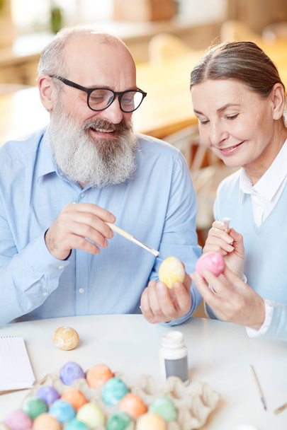 Aged bearded man with paintbrush pointing at one of Easter eggs while explaining his wife choice of color - Photo, Image