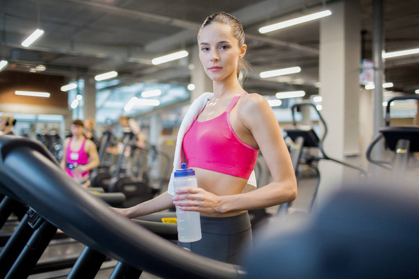 Young fit woman with plastic bottle of water looking at camera while standing by treadmill - Photo, Image