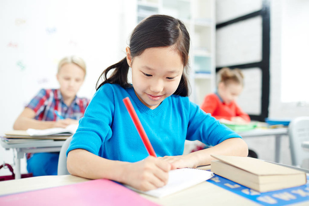 Asiática estudiante de primaria con dibujo de lápiz rojo en hoja de papel en la lección con compañeros de clase en el fondo
 - Foto, imagen