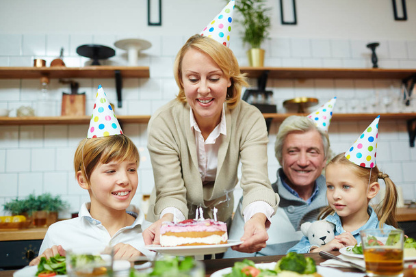 glückliche Frau stellt Geburtstagstorte auf festlichen Tisch für ihre Familie - Foto, Bild