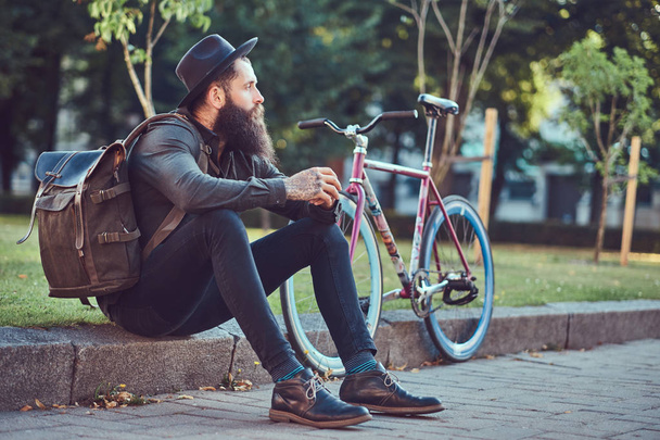Guapo viajero hipster con una elegante barba y tatuaje en sus brazos vestido con ropa casual y sombrero con una bolsa, sentado en la acera, descansando después de un paseo en bicicleta
. - Foto, imagen