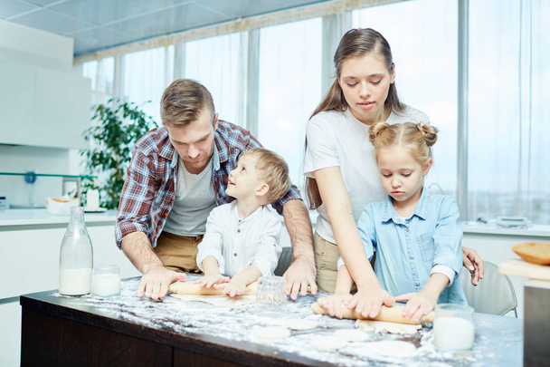Padre, madre, hija e hijo rodando masa para pasteles caseros en la mesa
 - Foto, imagen