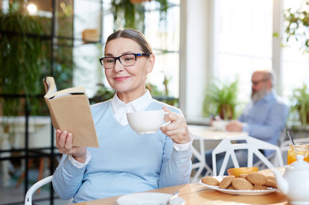 Contemporary senior woman having tea with cookies in cafe and reading book - Valokuva, kuva