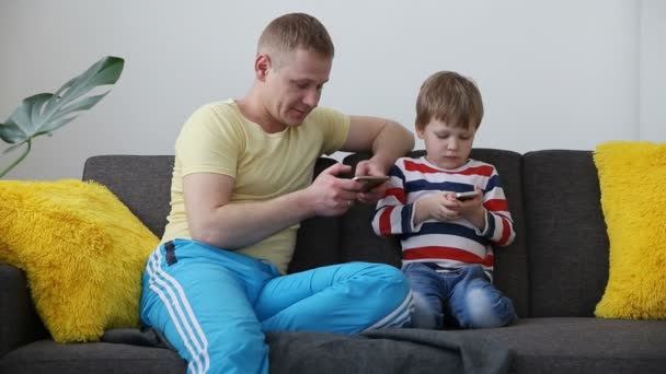Geräte im Leben einer modernen Familie. Vater mit kleinem Sohn spielt Spiele auf Smartphones, sitzt zu Hause auf dem Sofa. - Filmmaterial, Video