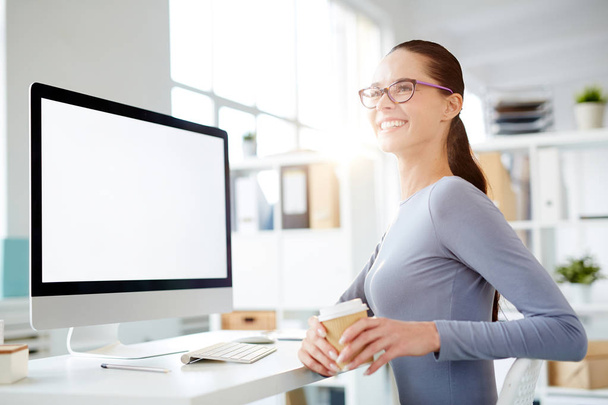 Счастливая молодая деловая женщина с выпивкой сидит на рабочем месте и смотрит на коллегу
 - Фото, изображение