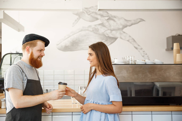 Coffee Business Concept - young smart bearded bartender enjoy talking and giving take away cup of coffee to pretty customer. - Photo, Image