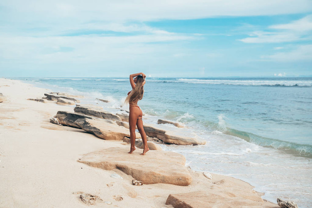 ΠΙΣΩ ΟΨΗ σέξι τατουάζ κορίτσι με κόκκινο μαγιό κρατά τα μαλλιά με τα χέρια να ποζάρει στην παραλία. Όμορφη ξανθιά γυναίκα χαλαρώνοντας στην παραλία στον ωκεανό. Έννοια των σπορ μοντέλο, μαγιό - Φωτογραφία, εικόνα