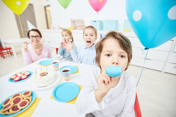 Niño pequeño con silbato de cumpleaños sentado junto a la mesa servida con sus amigos y maestro en el fondo
 - Foto, Imagen