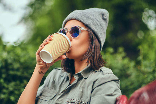 都市公園のベンチに座ってコーヒーを飲みながら帽子の流行に敏感な女性。自然でリラックス. - 写真・画像