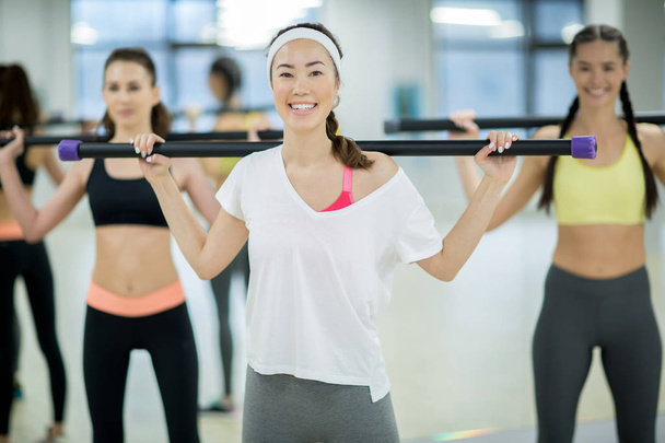 Happy active woman with training bar and her friends on background exercising in gym at leisure - Photo, Image