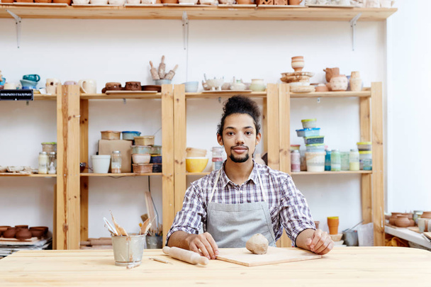 Joven artesano sentado por el lugar de trabajo en su taller con trozo de arcilla sobre tabla de madera y rodillo cerca
 - Foto, Imagen