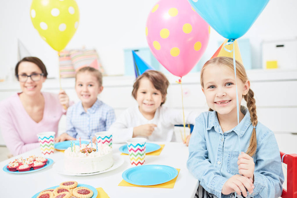 Grupy dzieci, wesoły i ich nauczyciel w urodziny czapki gospodarstwa balony siedząc przy stole podczas uroczystości - Zdjęcie, obraz