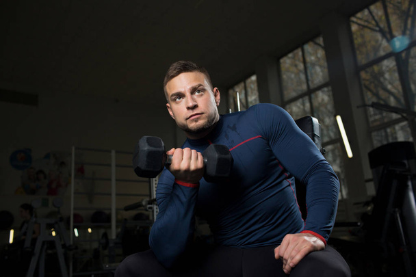 Young man holding dumbbell and exercising during workout in gym - Zdjęcie, obraz