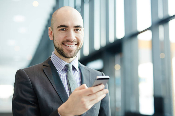 Kopasz fiatal üzletember toothy mosollyal nézett kamera, míg üzenet-ban smartphone - Fotó, kép