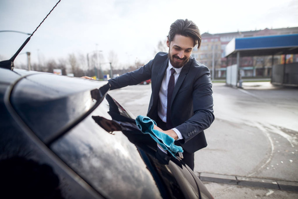 knappe stijlvolle jonge bebaarde man in pak auto met microfiber doek op de handmatige auto wassen self-service station reinigen - Foto, afbeelding
