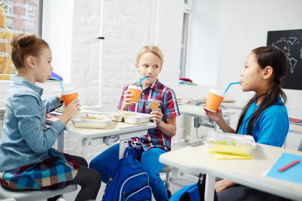 Три одноклассника в начальной школе обедают за партами и пьют из пластиковых стаканов
 - Фото, изображение