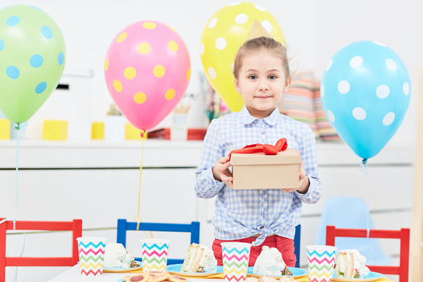Bambina con confezione regalo che guarda la macchina fotografica in piedi accanto al tavolo festivo con bevande e torte
 - Foto, immagini