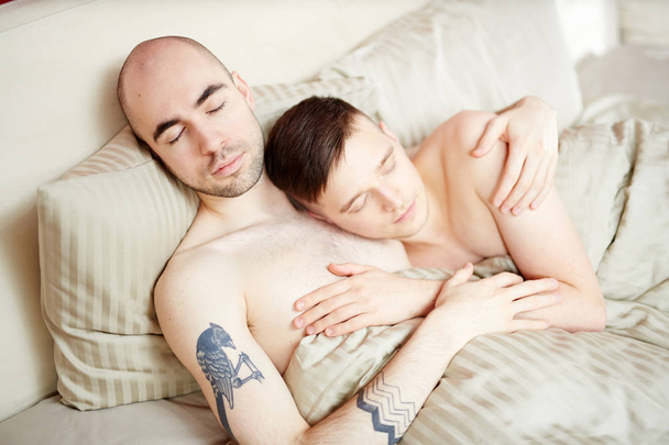 Молодые гомосексуальные любовники спят в постели под одеялом в объятиях
 - Фото, изображение