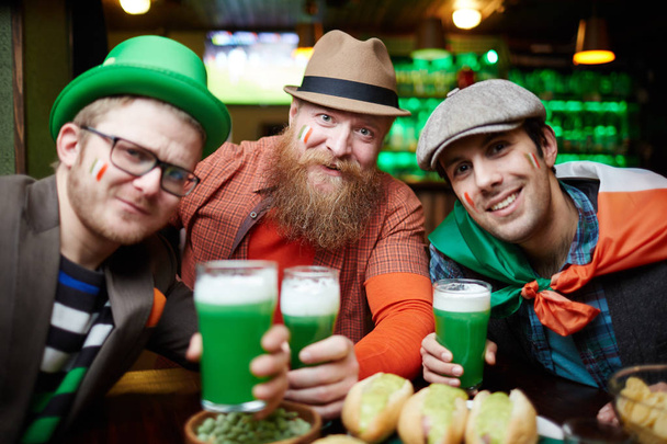 Grupo de hombres amigables brindando con usted mientras mira a la cámara durante la celebración de la fiesta nacional irlandesa
 - Foto, imagen