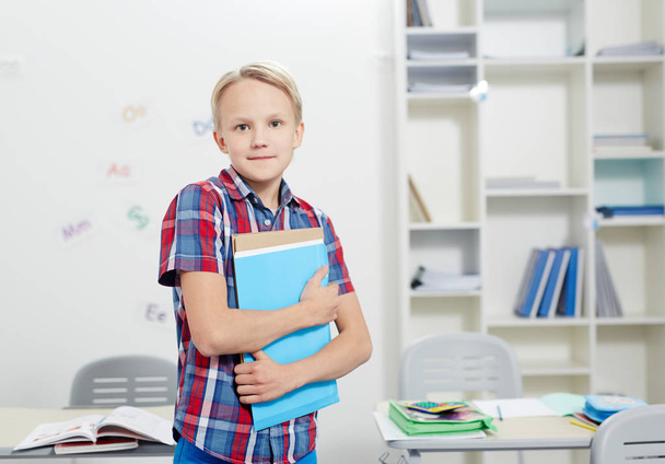 Diligent elementary schoolboy with copybooks looking at camera in classroom - Photo, Image
