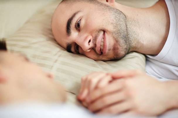 Χαμογελαστός νεαρός με το κεφάλι στο μαξιλάρι που βλέπουν τη σύντροφό του στο κρεβάτι - Φωτογραφία, εικόνα
