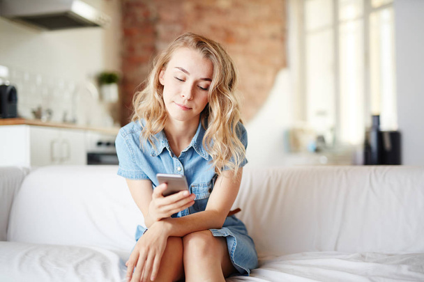 Genç kadın evde kanepede oturan ve onun smartphone içinde mesajlaşma - Fotoğraf, Görsel