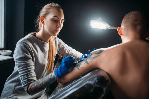 female tattooist making tattoo by machine on male shoulder, professional tattooing in salon - Photo, Image