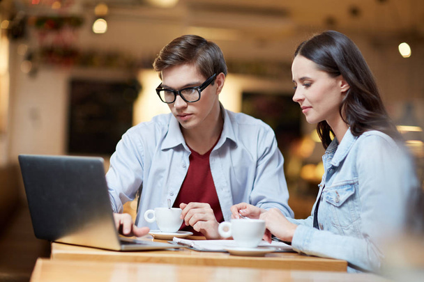 Dos jóvenes colegas con portátil viendo webinar mientras están sentados en la cafetería
 - Foto, Imagen