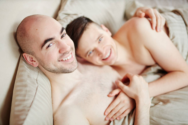 Joven cariñoso sonriendo gay los hombres acostado en la cama y mirando a la cámara
 - Foto, Imagen