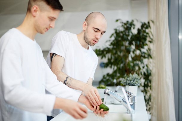 Δύο νεαροί ομοφυλόφιλοι, άνδρες πλύσιμο αβοκάντο στην κουζίνα στο σπίτι - Φωτογραφία, εικόνα