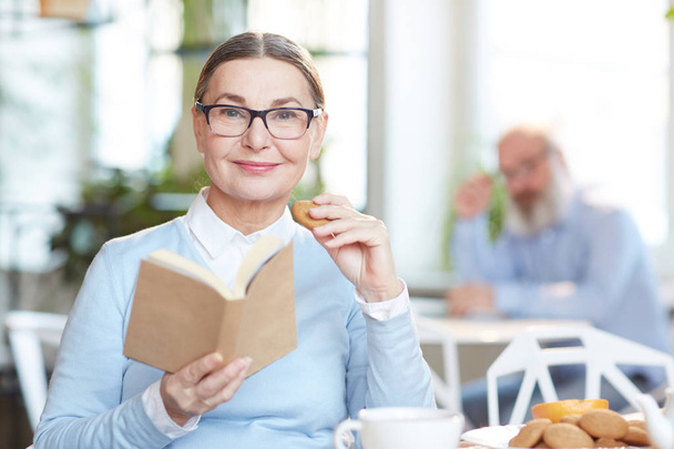 Mujer mayor en anteojos y ropa casual sentada en la cafetería con libro interesante y leyéndolo mientras toma galletas frescas
 - Foto, Imagen
