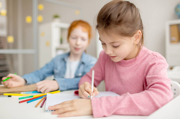Diligente chica con dibujo de crayón en la lección mientras su compañero de clase mirando su bloc de notas
 - Foto, imagen