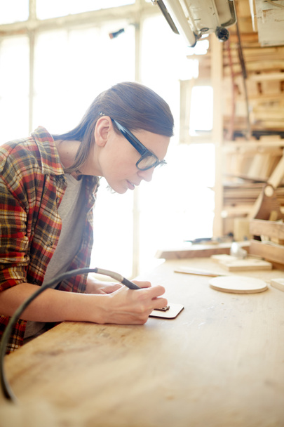 Joven carpintera femenina con equipamiento pirográfico dibujando ornamentos sobre tabla de madera
 - Foto, imagen