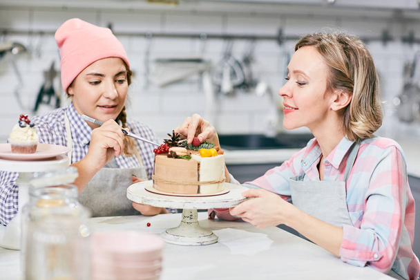 Dos mujeres jóvenes que decoran sabroso pastel festivo con frutas y bayas exóticas
 - Foto, Imagen