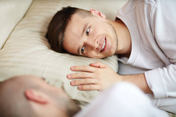 glücklich und verliebt junger schwuler Mann liegt auf Kissen und sieht seine Geliebte lächelnd an - Foto, Bild