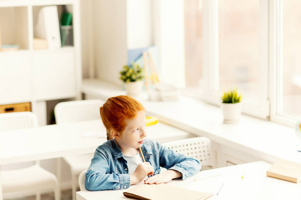 Задумчивая маленькая девочка сидит за столом, смотрит в окно класса и думает о новых идеях
 - Фото, изображение