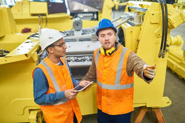 Two young machine operators wearing hardhats and reflective vests gathered together at production department of modern plant and trying to solve faced issue, one of them holding digital tablet - Photo, Image