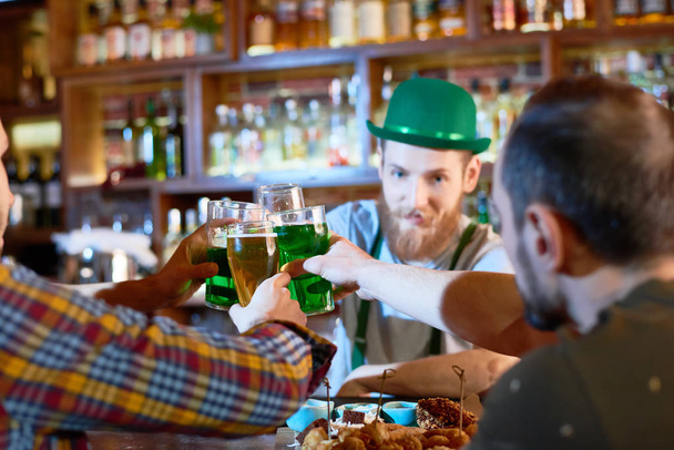 Cheerful young men toasting with beer glasses while hanging out in modern pub, handsome bearded barman wearing green bowler hat - Photo, Image