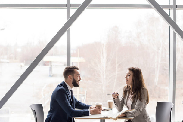 Retrato vista lateral de dos personas de negocios alegres, hombre y mujer, tomando café en la mesa en la cafetería sentado en la ventana grande
 - Foto, imagen