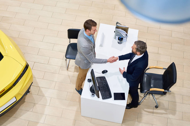 Above view of handsome salesman shaking hands with customer over table after closing successful car deal in dealership shop - Photo, Image