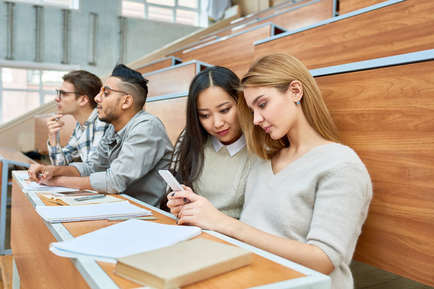 Międzynarodowej grupy studentów siedzi w wierszu na biurka w nowoczesne Aula czeka na klasy w nowoczesne college, skupić się na dwa ładne dziewczyny za pomocą smartfona - Zdjęcie, obraz