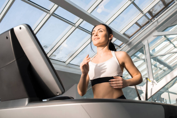 Waist up portrait of fit young woman enjoying running on treadmill in modern gym under glass roof - Фото, зображення