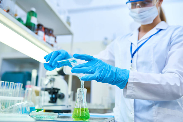 Portret van vrouwelijke wetenschapper het dragen van beschermende handschoenen gieten gekleurde vloeistoffen buizen testen tijdens de studie van chemische stoffen in medisch laboratorium te richten op handen - Foto, afbeelding