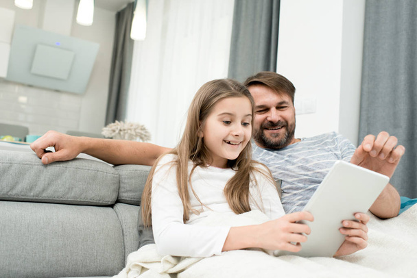 Бородатый мужчина средних лет и его маленькая дочь веселятся вместе, сидя на уютном диване и смотря мультики на цифровом планшете
 - Фото, изображение