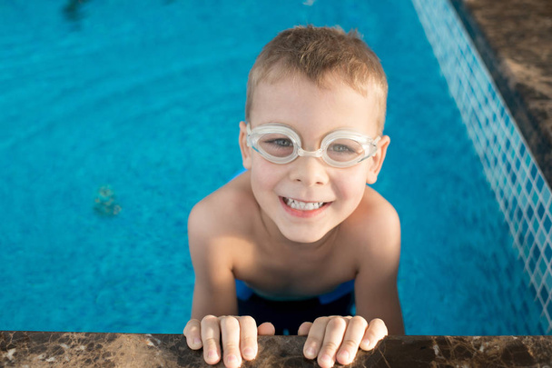 Κατακόρυφος βολή του χαριτωμένο μικρό αγόρι στο κολύμπι γυαλιά κοιτάζοντας τα φωτογραφικών μηχανών με toothy χαμόγελο, κρατώντας την άκρη της πισίνας - Φωτογραφία, εικόνα