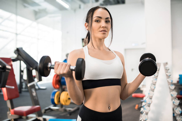 Portrait of sportive young woman  training with dumbbells in modern gym and looking at camera - Photo, Image