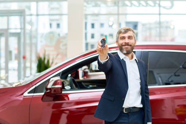 Ritratto di bell'uomo maturo che sorride felicemente alla macchina fotografica in piedi accanto alla nuovissima auto di lusso nello showroom e presenta le chiavi della macchina
 - Foto, immagini