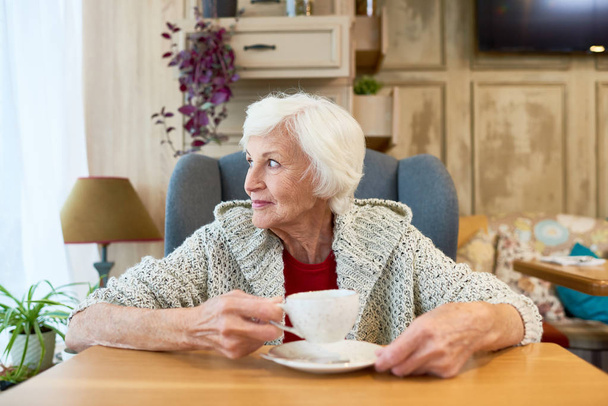 Талия портрет красивой пожилой женщины в трикотажном кардигане, наслаждающейся живописным видом из окна и согревающейся чашкой кофе, сидя в уютном маленьком кафе
. - Фото, изображение