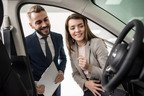 Portrait of cheerful young woman looking inside car while choosing luxury car in dealership showroom standing next to handsome salesman - Photo, Image