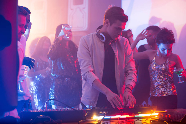 Porträt eines hübschen jungen DJs, der am Mischpult steht und während einer nächtlichen Party im Club Musik macht, Szene beleuchtet von Bühnenlichtern mit schönen Mädchen, die glitzernde Oberteile tragen und im Hintergrund tanzen - Foto, Bild